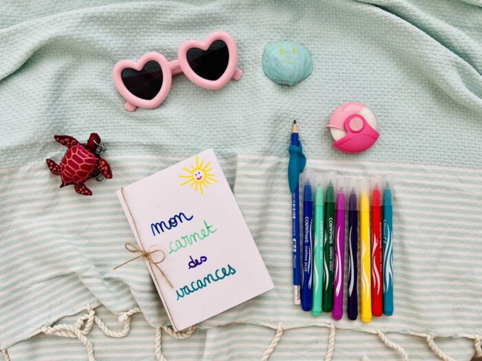 Le matériel du DIY carnet de souvenirs et d'émotions : feutres Color'Peps, crayon et taille-crayon Maped autour du carnet fait maison, sur un plaid de plage